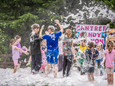 An image of the foam canon shooting out white foam which is covering the children standing underneath it and creating a blanket of sudds on the floor.